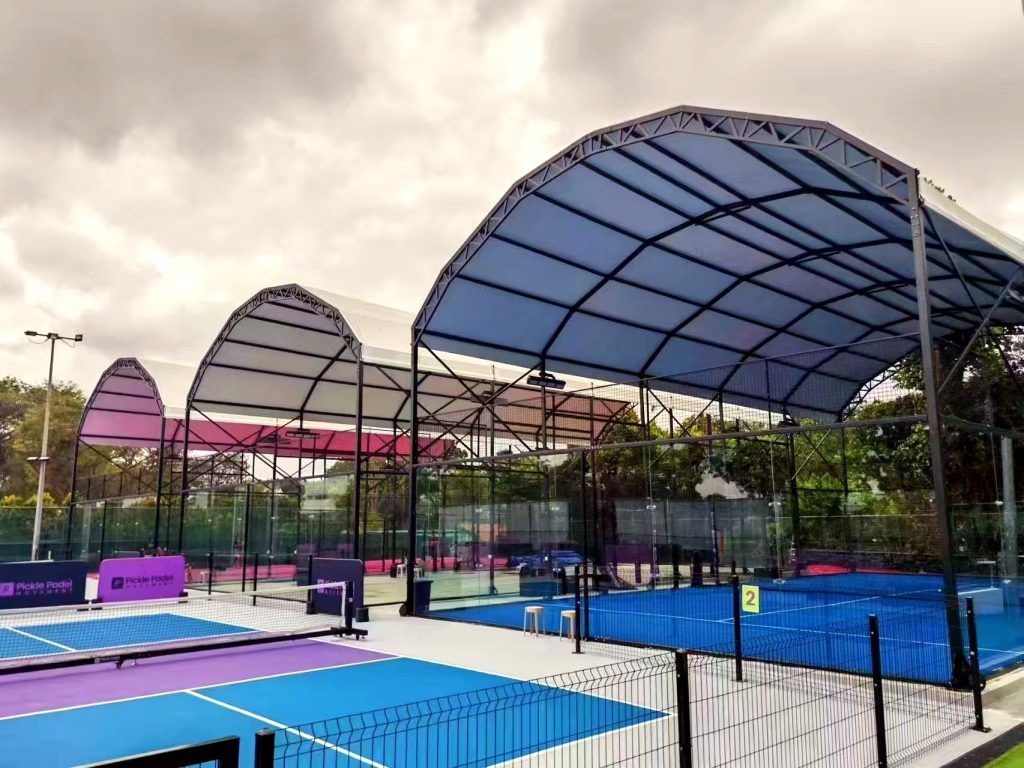 Covered padel court with curved steel roof and blue courts at an outdoor club